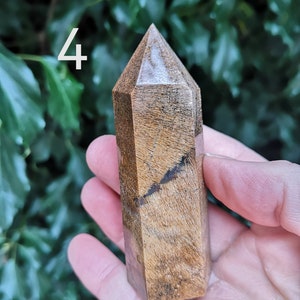 Puede incluir: Una punta de cristal marr&oacute;n y blanco con una parte superior puntiaguda y una superficie lisa y pulida. El cristal mide aproximadamente 10 cm de alto y se sostiene en una mano.