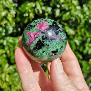 May include: A polished sphere of zoisite with ruby inclusions. The stone has a vibrant green base, speckled with black and accented by striking pink ruby clusters. The sphere is held in a hand against a backdrop of green foliage.