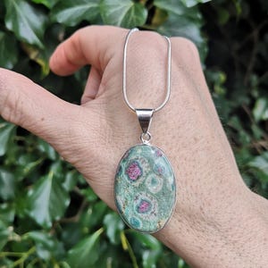 Peut inclure: Collier pendentif ovale avec une pierre précieuse polie sertie dans du métal argenté. La pierre affiche un motif vert et rose moucheté. La chaîne du collier est une fine chaîne serpent argentée.