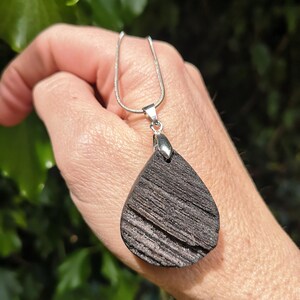 May include: A silver necklace featuring a teardrop-shaped pendant crafted from dark, textured wood. The pendant hangs from a fine silver chain. The background shows blurred green leaves.
