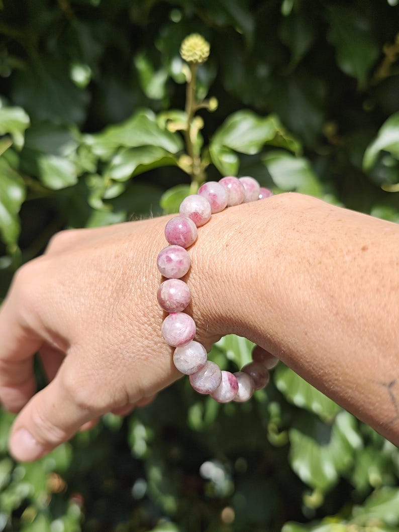 May include: A beaded bracelet featuring round, polished stones in varying shades of pink, magenta, and white. The beads are strung together in a continuous circular pattern, displaying natural marbling and translucent textures throughout each individual stone.