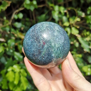 May include: A green and blue sphere with red flecks, polished to a smooth finish. The sphere is made of a natural stone, likely a type of jasper or agate.