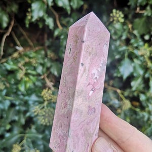 May include: A pink and white polished crystal point, also known as a wand or obelisk. The crystal is a natural stone with a smooth, polished surface.