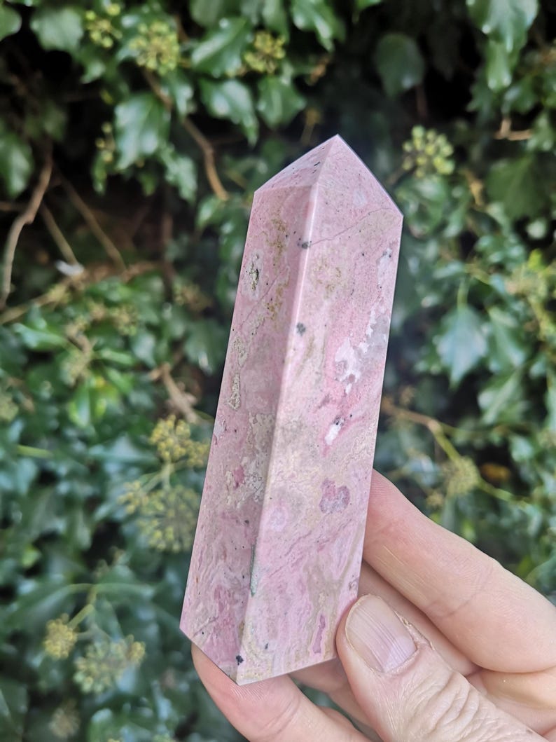 May include: A pink and white polished crystal point, also known as a wand or obelisk. The crystal is a natural stone with a smooth, polished surface.