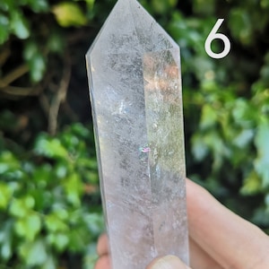 May include: A clear quartz crystal point with a slight iridescent sheen. The crystal is approximately 15 cm long and is held in a hand.