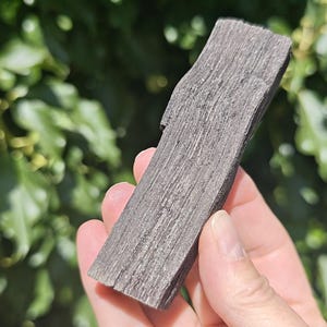 May include: A rectangular piece of dark grey, weathered wood with a prominent vertical grain and a rough, fibrous texture. The object has a slightly uneven surface with visible splintering along the edges and a dense, matte appearance throughout.