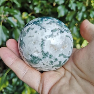May include: A green and white moss agate sphere, polished to a smooth finish. The sphere is held in a hand, showcasing its intricate patterns and natural beauty.
