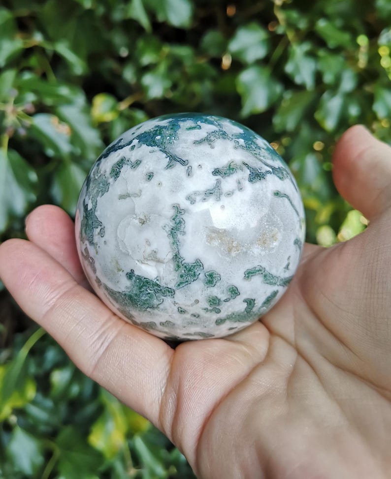 May include: A green and white moss agate sphere, polished to a smooth finish. The sphere is held in a hand, showcasing its intricate patterns and natural beauty.