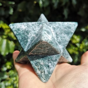 May include: A green and white stone Merkaba star. The geometric shape is held in a hand, with a natural green background. The stone has a speckled pattern and a polished finish. The Merkaba is a symbol of spiritual transformation.