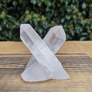 May include: A clear quartz crystal cluster with two points, forming an X shape. The crystal is sitting on a wooden surface.