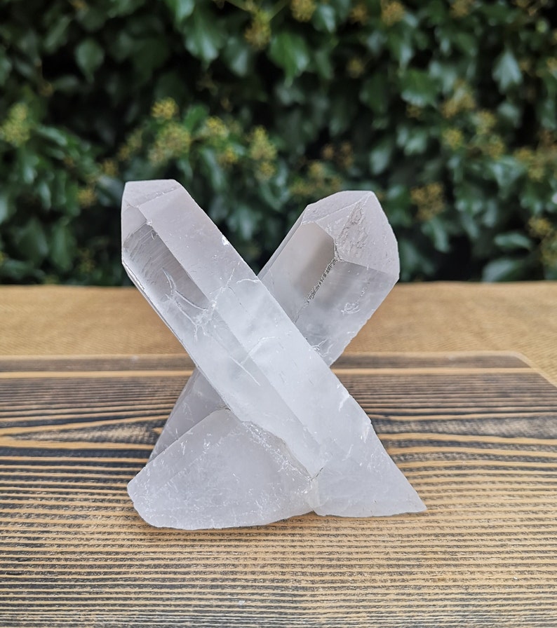 May include: A clear quartz crystal cluster with two points, forming an X shape. The crystal is sitting on a wooden surface.