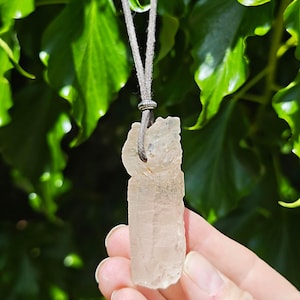 May include: A raw, light-coloured crystal pendant hanging from a grey cord. The crystal is rectangular with a rough, textured surface. The background is a blurred view of green foliage.