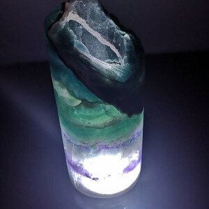 May include: A green, purple, and white fluorite crystal with a light shining through it. The crystal is shaped like a cylinder and has a rough, natural texture.