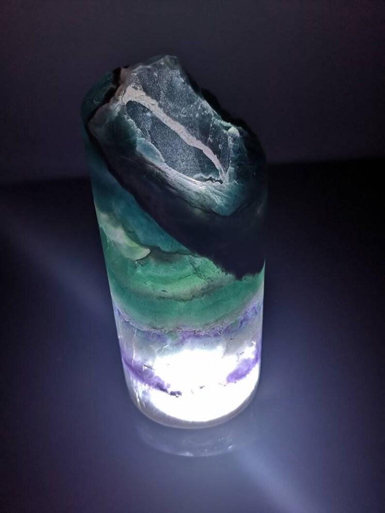 May include: A green, purple, and white fluorite crystal with a light shining through it. The crystal is shaped like a cylinder and has a rough, natural texture.
