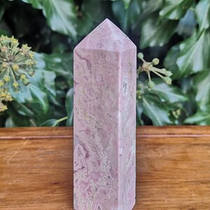 May include: A pink and white striped crystal point, also known as a crystal wand, is displayed on a wooden surface. The crystal point is approximately 12.5 cm tall.