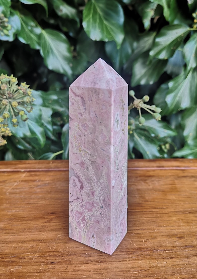 May include: A pink and white striped crystal point, also known as a crystal wand, is displayed on a wooden surface. The crystal point is approximately 12.5 cm tall.