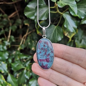 May include: Oval pendant necklace featuring a polished stone with a teal and magenta pattern, set in a silver-colored bezel. The pendant hangs from a delicate silver chain, showcasing a natural gemstone design.