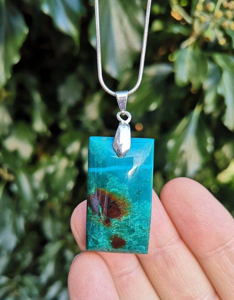 Pu&ograve; includere: Un ciondolo rettangolare con un vivace motivo turchese e marrone, sospeso a una catena d'argento. Il ciondolo ha un'ansa d'argento. Lo sfondo &egrave; una vista sfocata di fogliame verde.