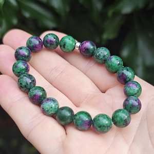 May include: A beaded stretch bracelet composed of round, polished zoisite stones featuring a mottled pattern of deep forest green and vibrant magenta-pink inclusions, accented by a single small, smooth, cylindrical silver-toned metal spacer bead.