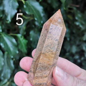 Puede incluir: Una punta de cristal marr&oacute;n y naranja con una punta puntiaguda y una forma facetada. El cristal se sostiene en una mano contra un fondo de hojas verdes. El n&uacute;mero "5" es visible en la esquina superior izquierda de la imagen.