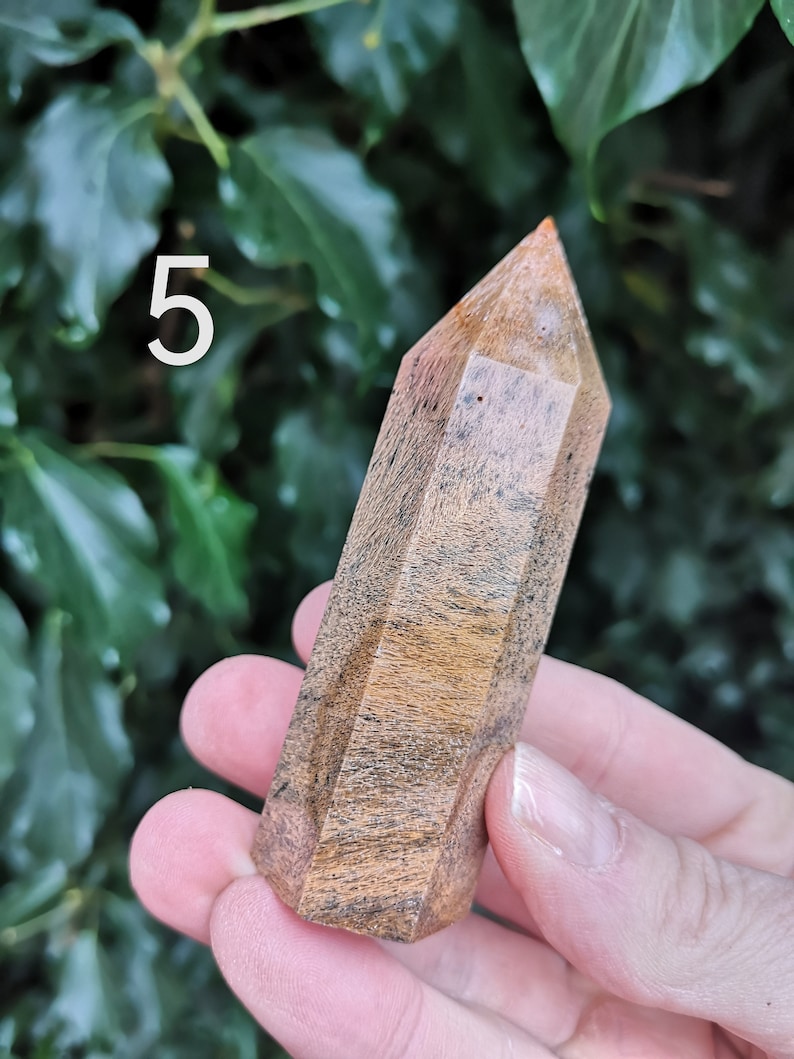 Puede incluir: Una punta de cristal marr&oacute;n y naranja con una punta puntiaguda y una forma facetada. El cristal se sostiene en una mano contra un fondo de hojas verdes. El n&uacute;mero "5" es visible en la esquina superior izquierda de la imagen.