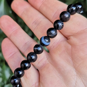 May include: A black beaded bracelet with white and gray circular patterns. The bracelet is made of polished, round beads. The beads are strung together to form a bracelet. The bracelet is held in a hand.