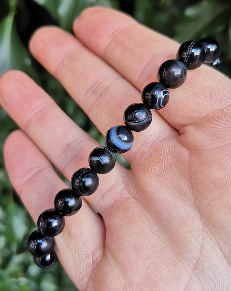 May include: A black beaded bracelet with white and gray circular patterns. The bracelet is made of polished, round beads. The beads are strung together to form a bracelet. The bracelet is held in a hand.