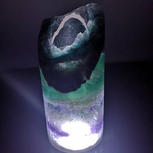 May include: A cylindrical, translucent, green, purple, and white fluorite crystal with a light inside. The crystal is illuminated from within, creating a soft glow.