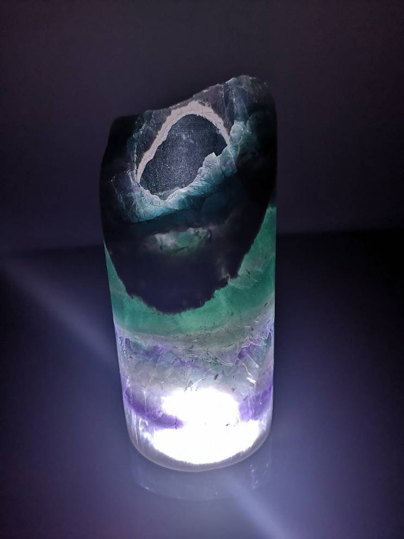 May include: A cylindrical, translucent, green, purple, and white fluorite crystal with a light inside. The crystal is illuminated from within, creating a soft glow.