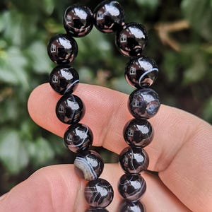 May include: A black beaded bracelet with a glossy finish. The beads are round and feature white and gray banding patterns. The bracelet is held up against a blurred green background.