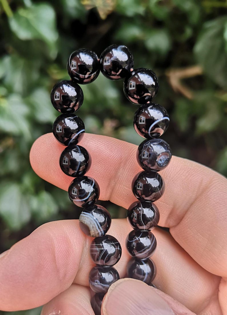May include: A black beaded bracelet with a glossy finish. The beads are round and feature white and gray banding patterns. The bracelet is held up against a blurred green background.