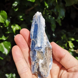 K&ouml;nnte beinhalten: Ein blauer Kyanit-Kristall mit silbernem Schimmer. Der Kristall ist oben spitz und hat eine raue, nat&uuml;rliche Textur.