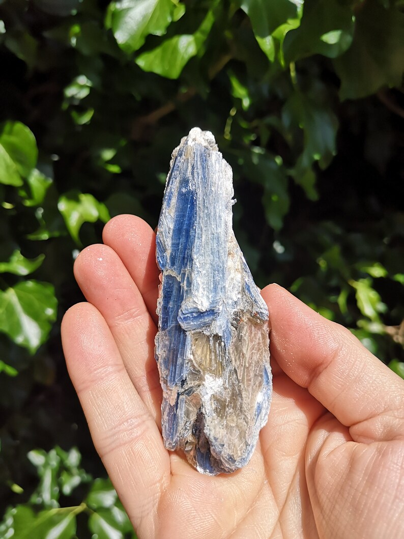 K&ouml;nnte beinhalten: Ein blauer Kyanit-Kristall mit silbernem Schimmer. Der Kristall ist oben spitz und hat eine raue, nat&uuml;rliche Textur.