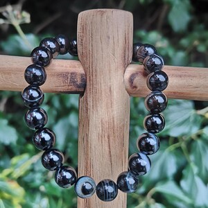 May include: A black beaded bracelet with a glossy finish. The beads are round and appear to be made of polished stone, with some beads showing a subtle white and gray pattern. The bracelet is displayed on a wooden stand against a backdrop of green foliage.