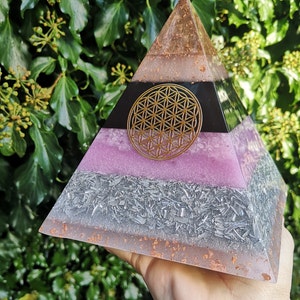 May include: A multi-layered pyramid made of resin with a flower of life design in gold on the top layer. The pyramid is pink, black, silver, and copper.