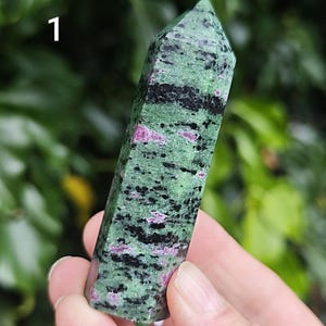 May include: A polished, hexagonal crystal tower featuring a mottled pattern of forest green, deep black, and vibrant magenta-pink inclusions. The stone has a pointed top and a smooth, faceted surface, with the number 1 visible in the upper left corner.