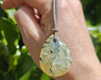 May include: A teardrop-shaped pendant necklace featuring a translucent, pale yellow stone with black inclusions. The stone is suspended from a gray cord. The background is blurred green foliage.