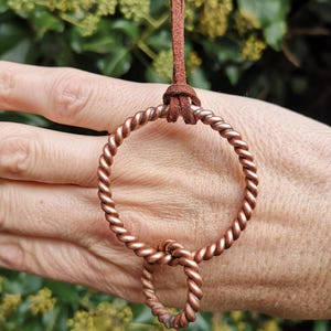 May include: A copper pendant necklace with two twisted metal rings, a large and a small one, joined by a loop. It hangs from a brown suede cord, knotted at the top. The background is blurred green foliage.