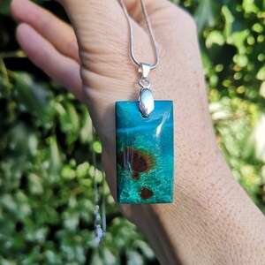 Pu&ograve; includere: Una collana con pendente rettangolare con catena argentata. Il pendente &egrave; di un vibrante colore verde acqua con inclusioni marroni e bianche. La collana &egrave; tenuta su uno sfondo di fogliame verde.