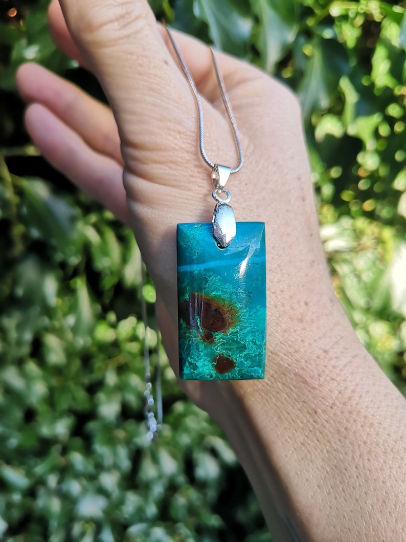 Pu&ograve; includere: Una collana con pendente rettangolare con catena argentata. Il pendente &egrave; di un vibrante colore verde acqua con inclusioni marroni e bianche. La collana &egrave; tenuta su uno sfondo di fogliame verde.