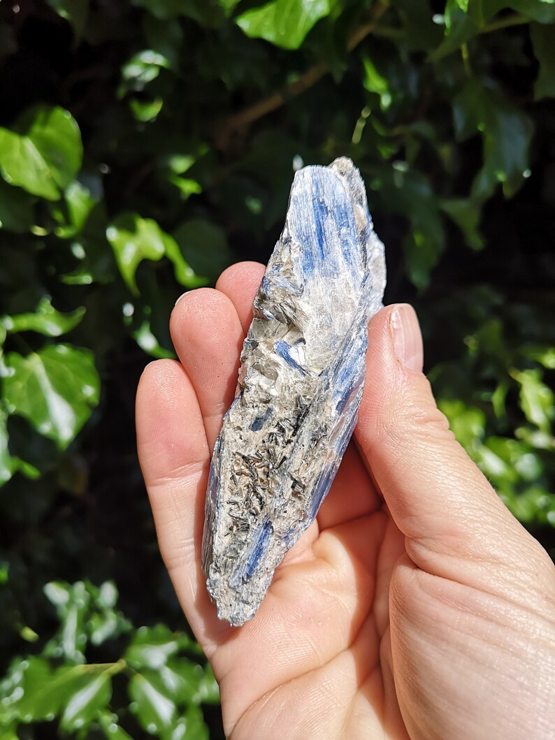 K&ouml;nnte beinhalten: Ein blauer Kyanit-Kristall mit rauer, nat&uuml;rlicher Textur. Der Kristall ist hellblau mit wei&szlig;en und grauen Streifen. Es ist ein roher, unpolierter Kristall.