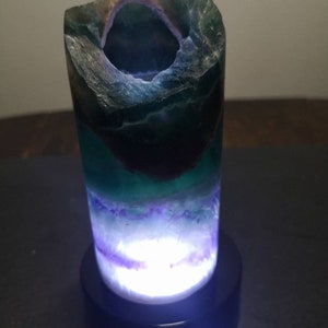 May include: A green, purple, and white fluorite crystal lamp with a black base. The crystal is hollowed out and lit from within, creating a glowing effect.