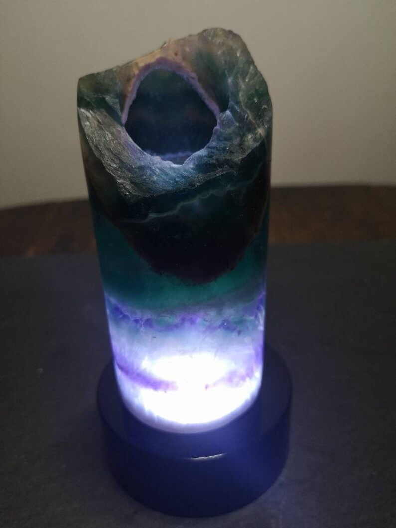 May include: A green, purple, and white fluorite crystal lamp with a black base. The crystal is hollowed out and lit from within, creating a glowing effect.