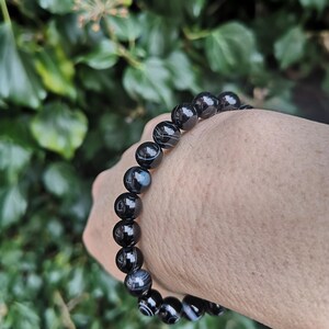 May include: A black and white striped bead bracelet. The beads are round and polished, with a glossy finish. The bracelet is strung on an elastic band. The background is a blurred view of green foliage.