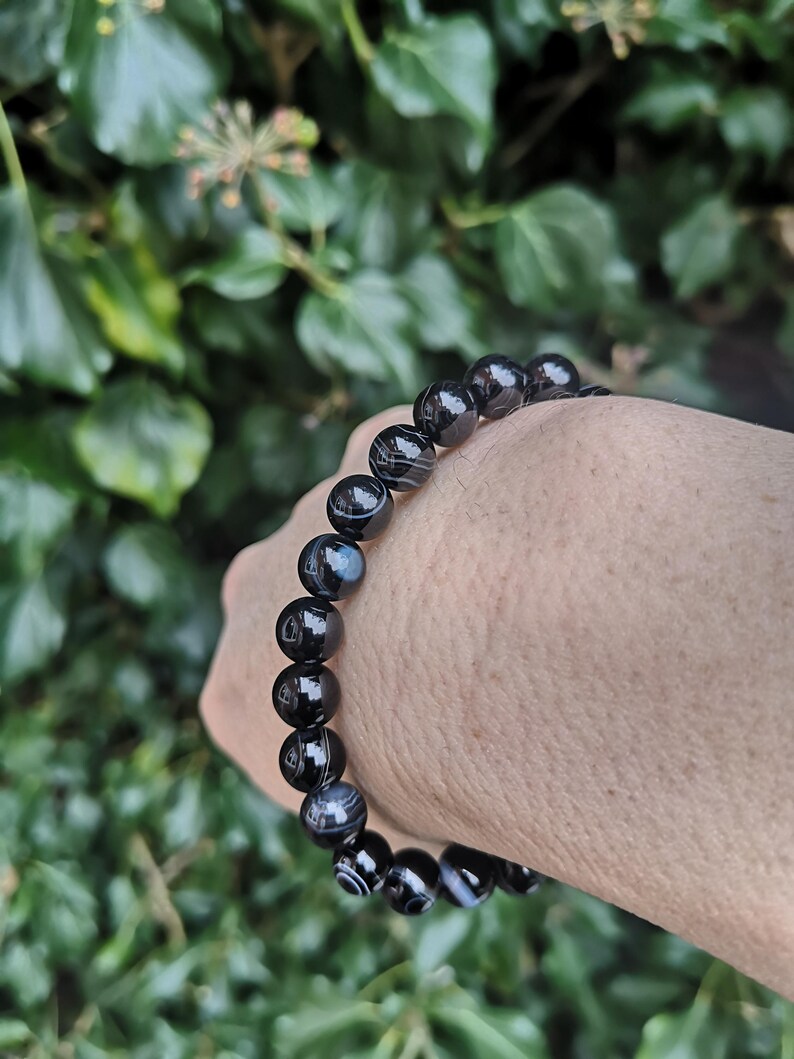 May include: A black and white striped bead bracelet. The beads are round and polished, with a glossy finish. The bracelet is strung on an elastic band. The background is a blurred view of green foliage.