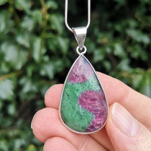 May include: A teardrop-shaped pendant necklace with a silver chain. The pendant features a polished stone with a mix of green and pink hues, set in a silver bezel. The necklace is held by a hand against a blurred green background.