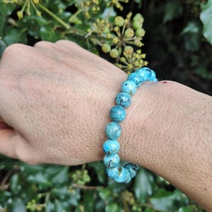 May include: A turquoise beaded bracelet with a stretch band. The round beads are a mottled blue-green colour. The bracelet is shown against a background of green foliage.