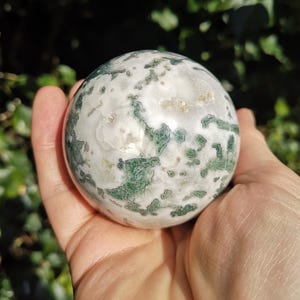 May include: A green and white agate sphere, polished to a smooth finish. The sphere has a swirling pattern of green and white, with a few small, clear crystals embedded in the surface.