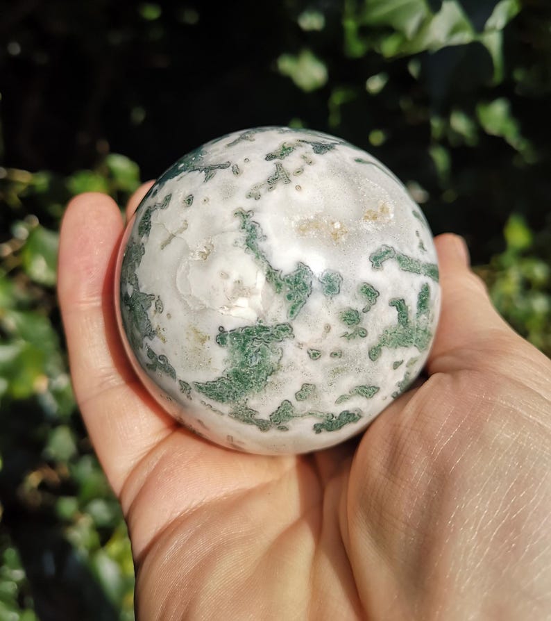 May include: A green and white agate sphere, polished to a smooth finish. The sphere has a swirling pattern of green and white, with a few small, clear crystals embedded in the surface.