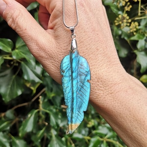 May include: A pendant necklace with a large, iridescent blue and teal feather-shaped stone. The stone has a silver-coloured metal cap and hangs from a delicate silver chain. The feather has a gold-coloured tip and detailed veining.